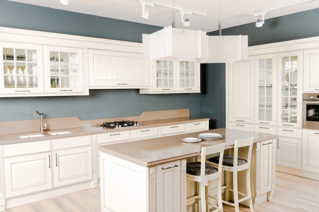 A modern kitchen with white cabinetry and beige countertops features premium kitchen installations, including a built-in oven, a gas stovetop, a stainless steel sink, and an island with two bar stools. The walls are painted in a dark grayish-blue color, illuminated by hanging lights above.