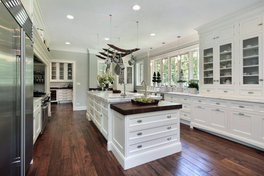 A spacious, modern kitchen features two large islands with dark wood countertops, white cabinetry with glass fronts, stainless steel appliances, and hardwood floors. Natural light streams in through tall windows, illuminating the clean, bright space—a perfect example of top-tier kitchen installations.