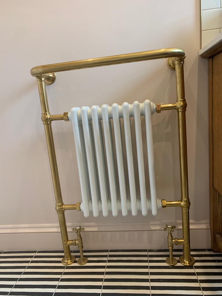 A white radiator with gold brass piping and fittings stands against a light-colored wall, positioned on a black-and-white striped tile floor next to a wooden cabinet.
