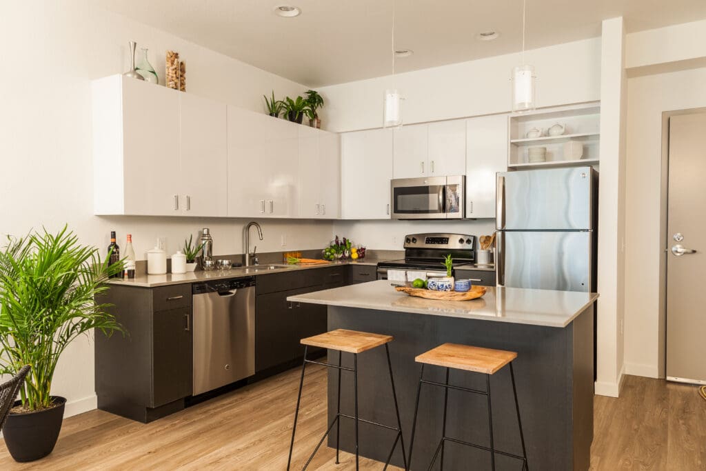 Modern kitchen installations feature white upper cabinets, black lower cabinets, stainless steel appliances, and light wood flooring. An island with two wooden stools sits at the center, while green plants add a fresh touch.