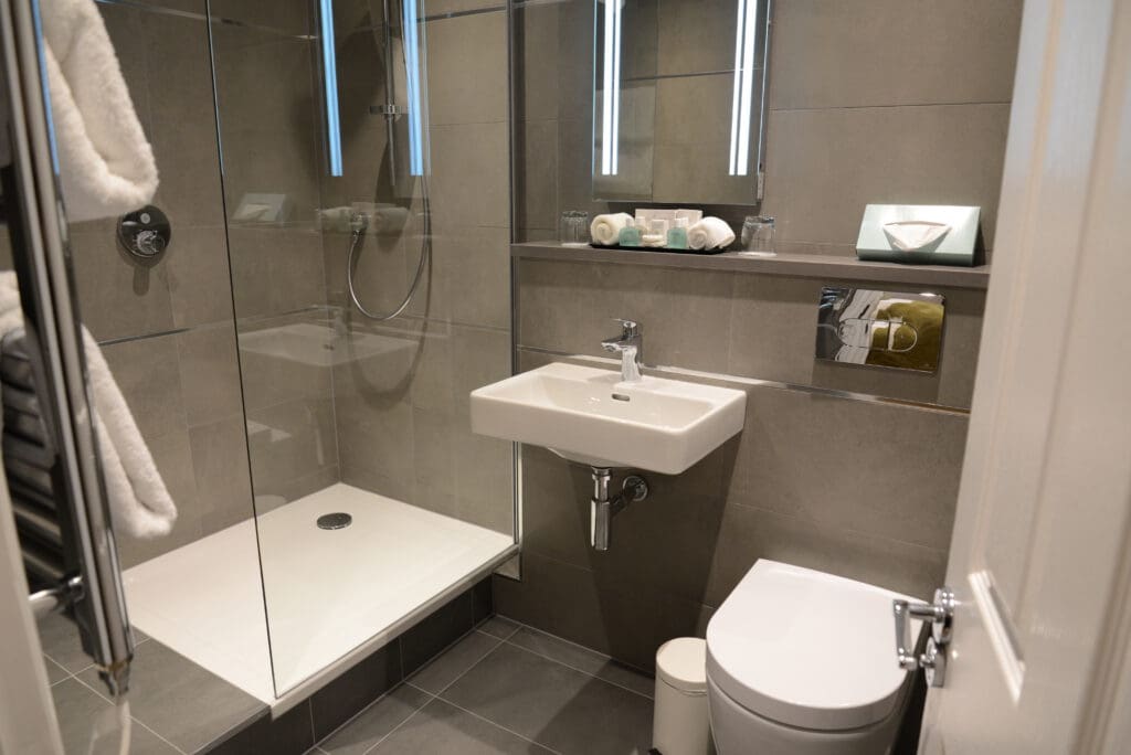 Modern bathroom featuring updated bathroom fixtures, including a glass-enclosed shower, square white sink, and wall-mounted toilet. Towels and toiletries are neatly arranged on shelves, with gray-toned tiles enhancing the fresh look.