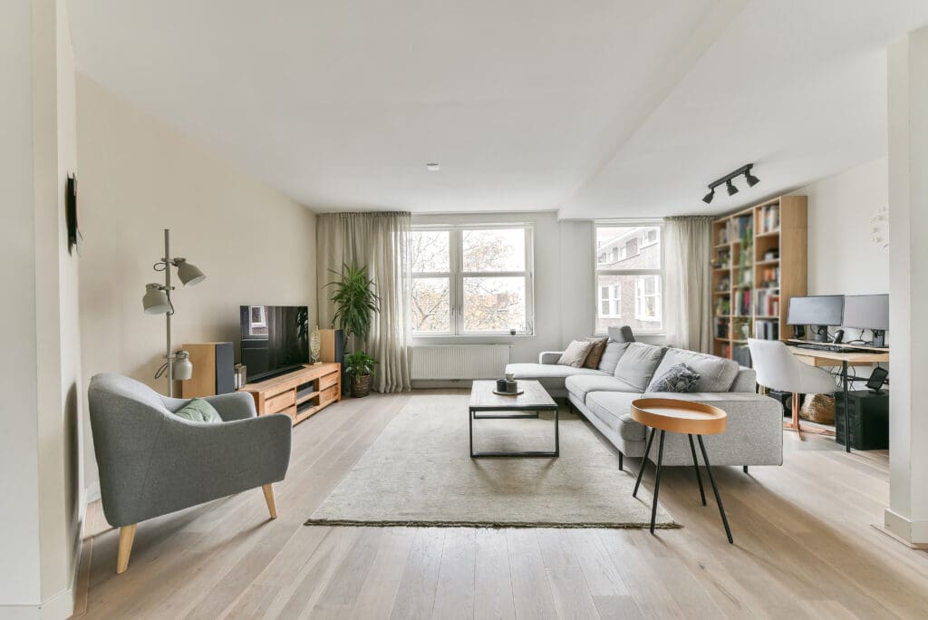 Bright, modern living room with light wood floors, a gray sofa, armchair, coffee table, TV on a wooden stand, large windows, indoor plant, rug, bookshelves, and a computer desk in the corner.
