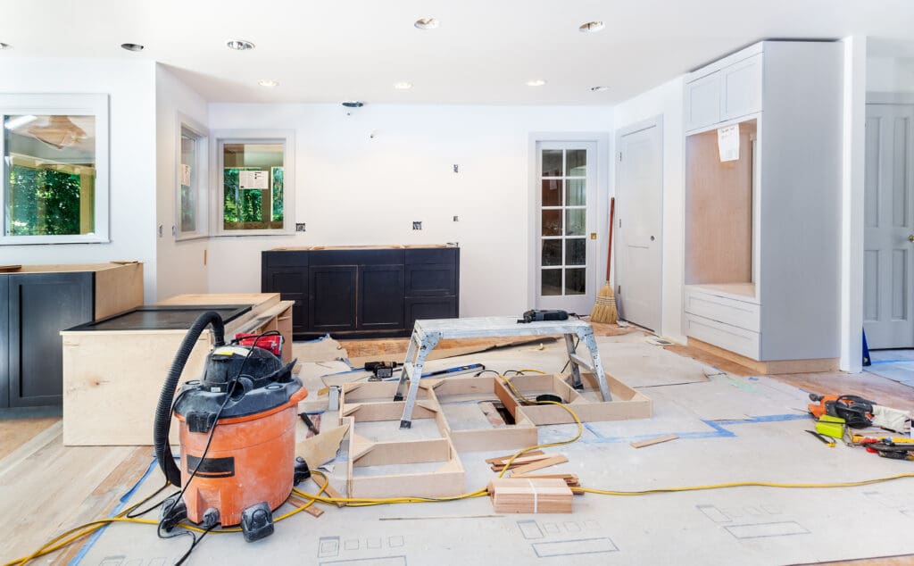 A kitchen under construction highlights the remodeling process, with cabinets being installed, tools, a vacuum, and materials scattered on the floor. Unfinished woodwork and partially painted walls showcase ongoing home renovation efforts.