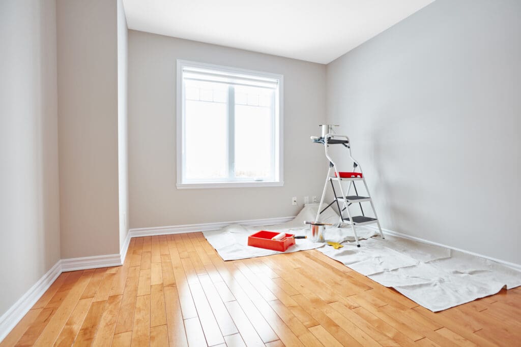 A bright, empty room with light wood floors and gray walls. Natural light pours in through a window. A step ladder, paint tray, roller, and drop cloths suggest ongoing home renovation or property remodeling.