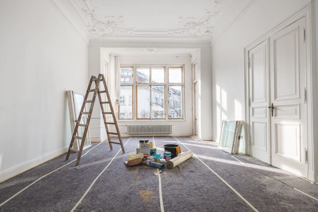 A bright, empty room undergoing home renovation with a ladder, paint cans, and tools scattered on protective sheets. Sunlight streams in through large windows as the space transforms during property remodeling.