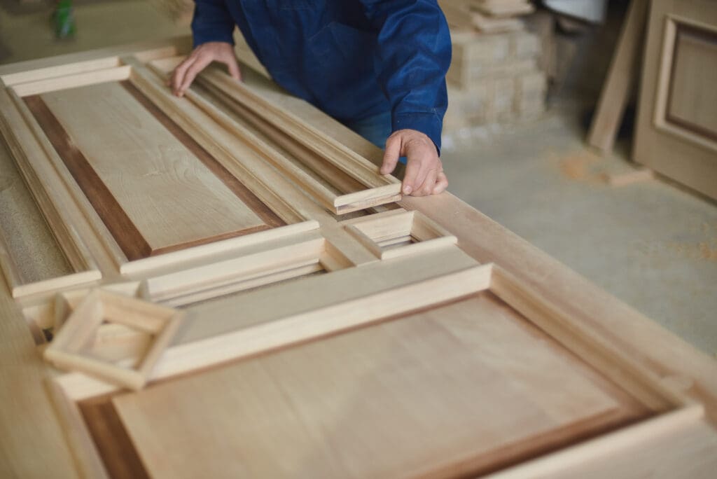 A carpenter in a blue shirt assembles wooden panels and frames on a workbench in a carpentry workshop, carefully fitting a rectangular piece into place among other unfinished wood parts as part of quality carpentry services.