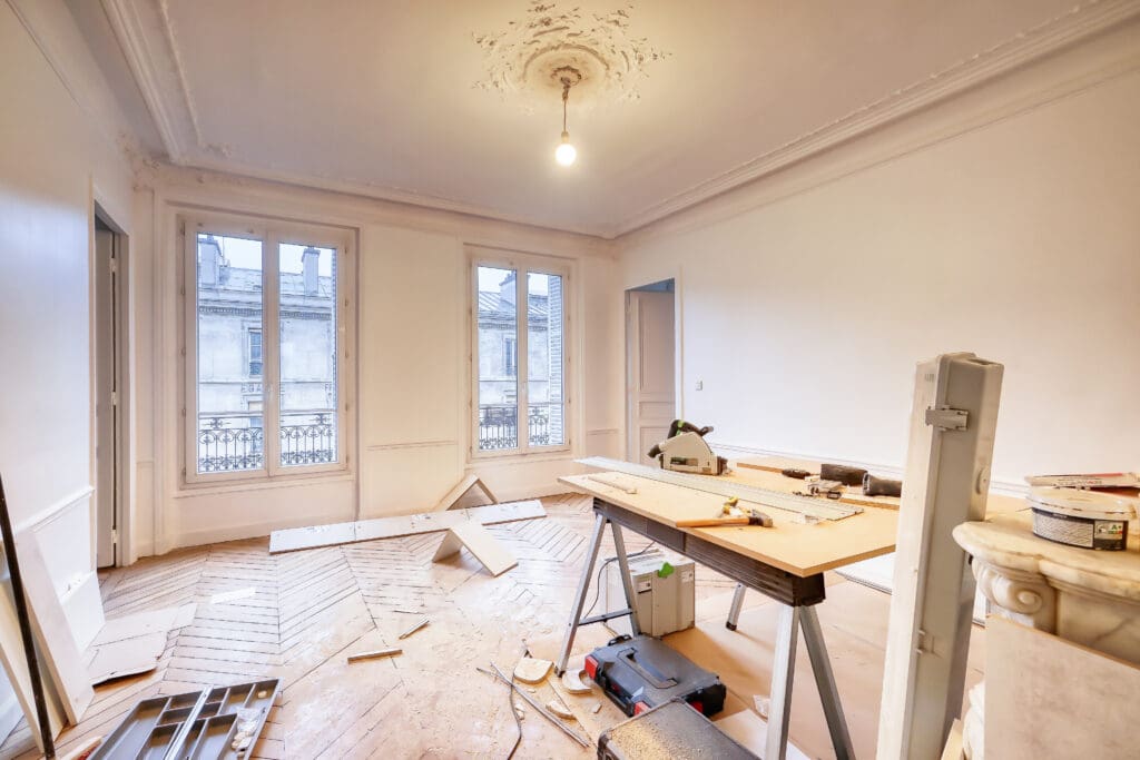 A bright room undergoing home renovation, with wooden parquet flooring partially installed, tools and materials scattered, a workbench in the center, and large windows letting in natural light.