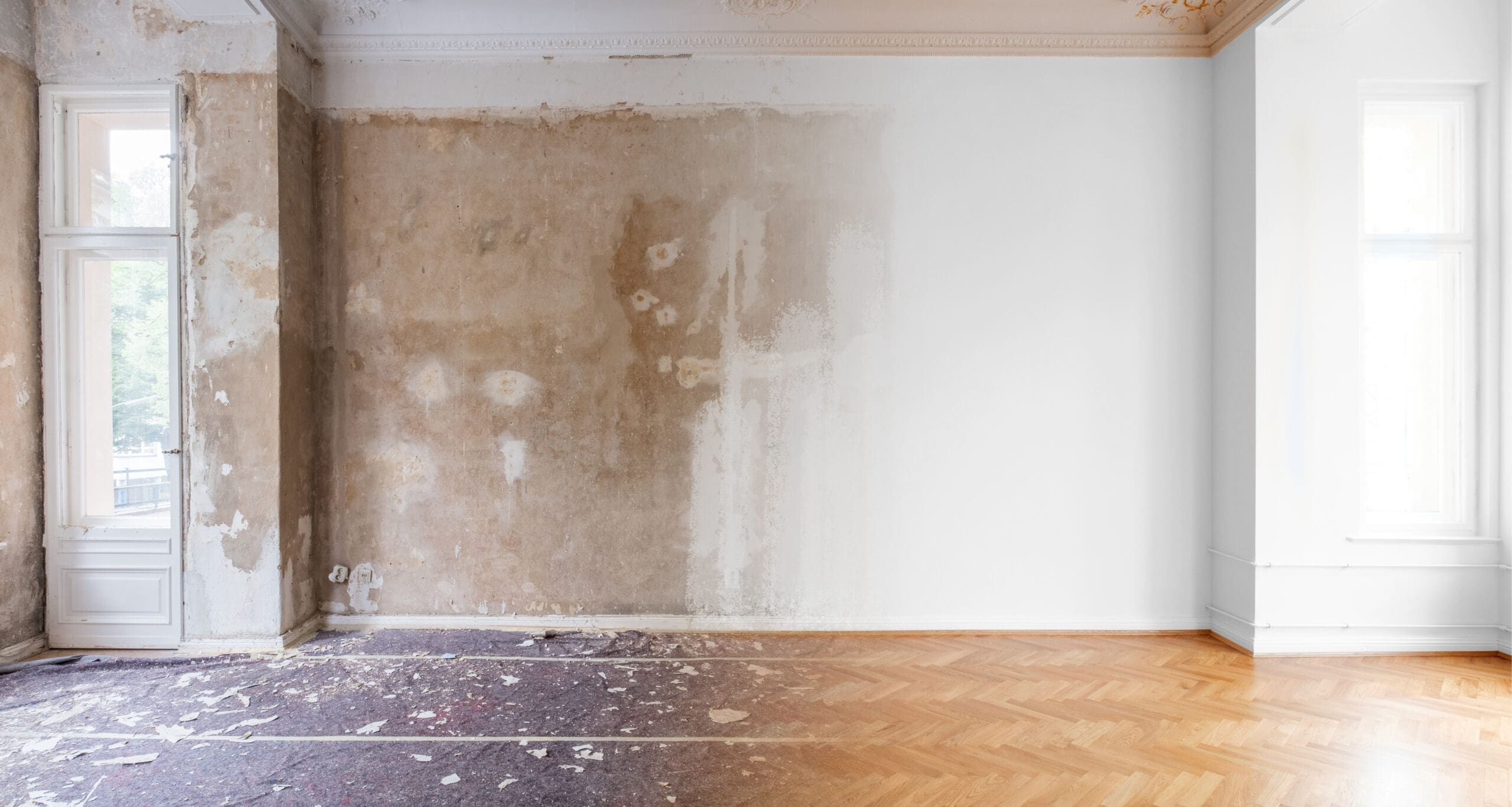 A partially renovated room with bare plaster on one wall, remnants of old paint, exposed wooden flooring, and two tall windows letting in natural light—a snapshot of home renovations in progress, where one side is unfinished and the other freshly painted.