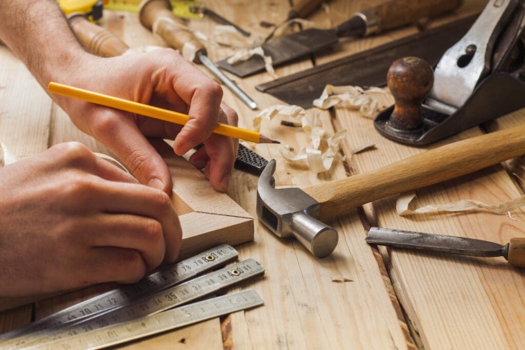 A person marks a piece of wood with a pencil on a workbench, surrounded by woodworking tools like a hammer, chisel, folding ruler, and wood shavings—perfect for carpenters or anyone interested in carpentry services.