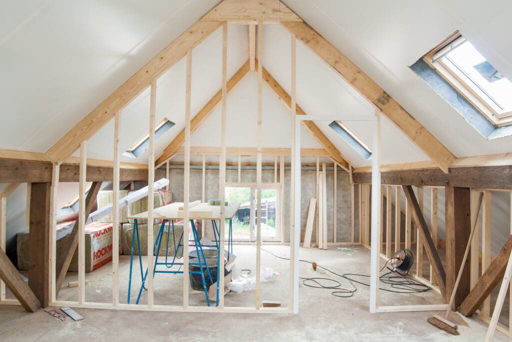 An unfinished attic renovation with exposed wooden framing, insulation materials, and tools scattered on the floor—a clear snapshot of home remodeling in progress. Sunlight streams in through skylight windows set into the slanted ceiling.