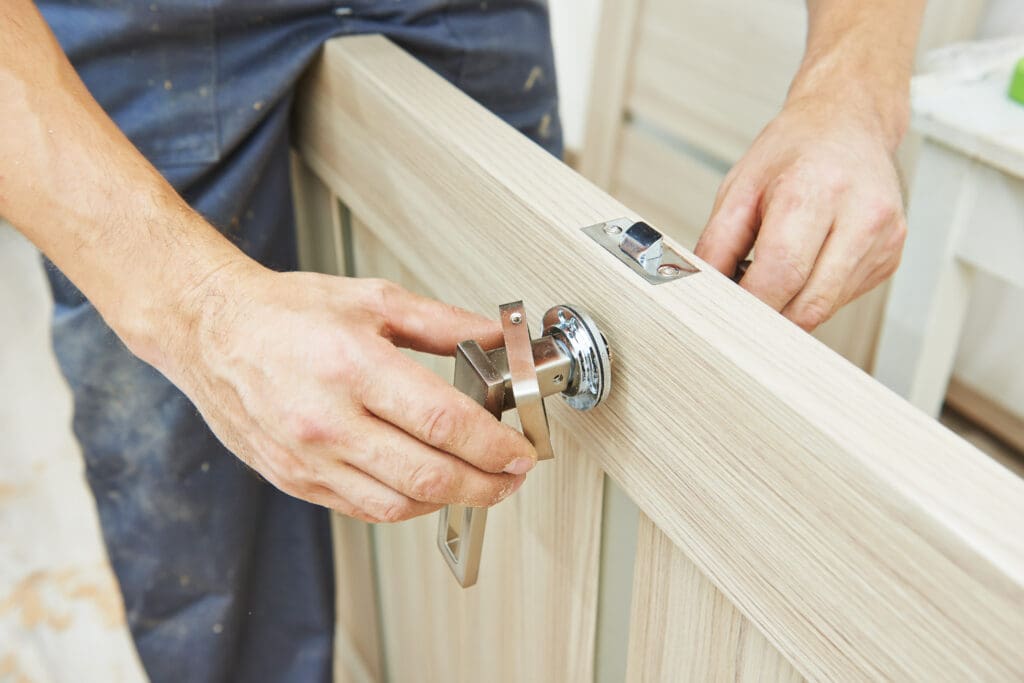 A carpenter installs a silver door handle and lock mechanism onto a light wood door, using both hands to fit the hardware in place.