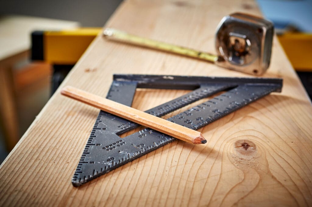 A carpenter’s pencil, a metal triangular ruler, and a tape measure rest on a wooden plank, suggesting skilled carpenters at work or carpentry services in progress.