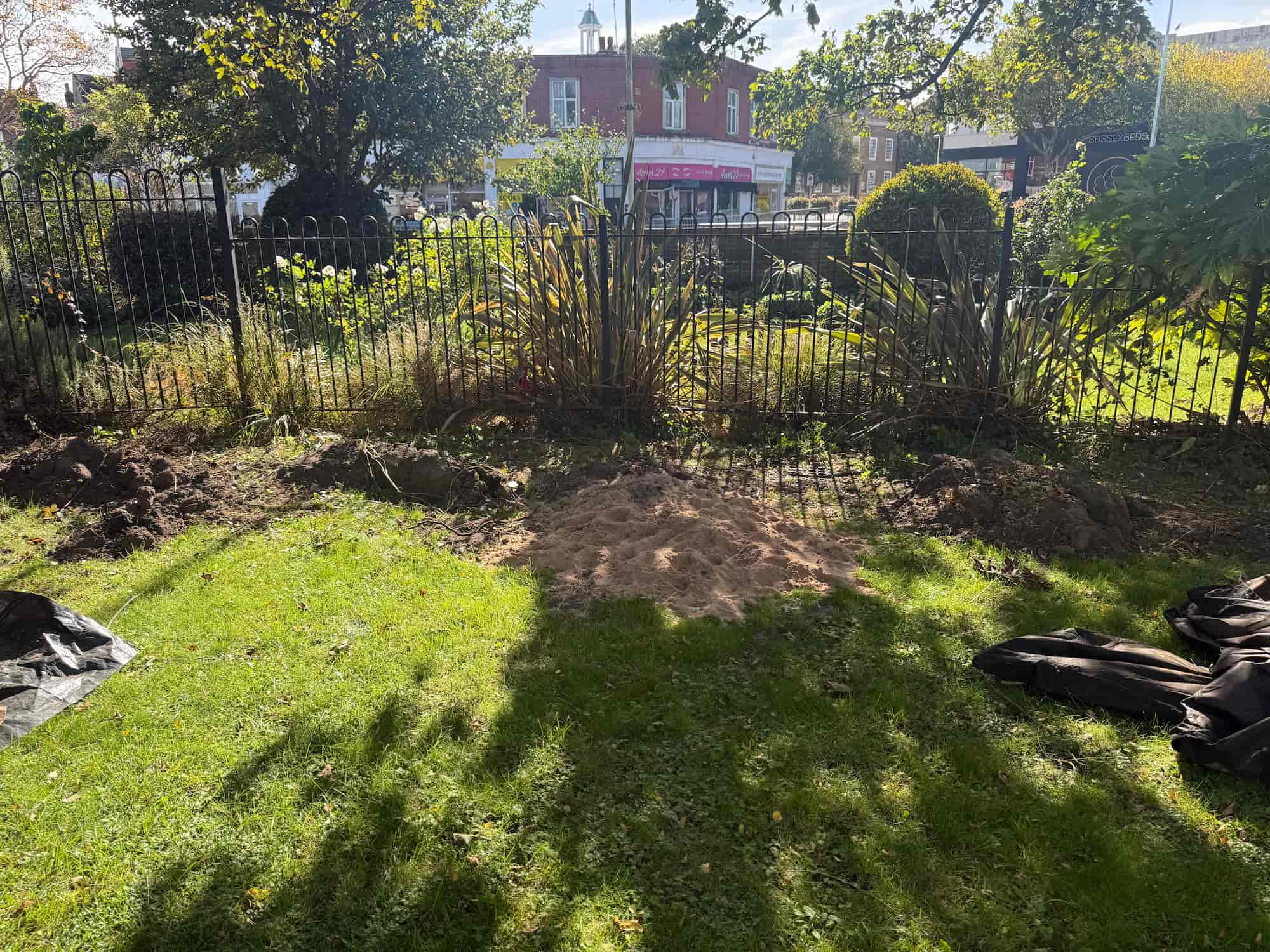 A patch of dug-up soil and sand is in front of a black metal fence in a park, with plants and shrubs along the fence. Sunlight and tree shadows cover parts of the green grass. Buildings are visible in the background.