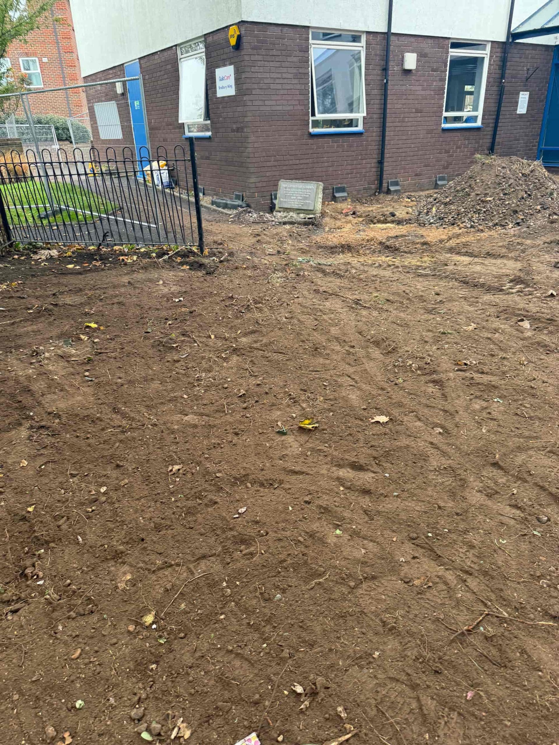 A patch of bare, freshly cleared earth in front of a brick building with white upper walls, two windows, a black fence, and a sign by the entrance. There are piles of dirt and some scattered leaves on the ground.