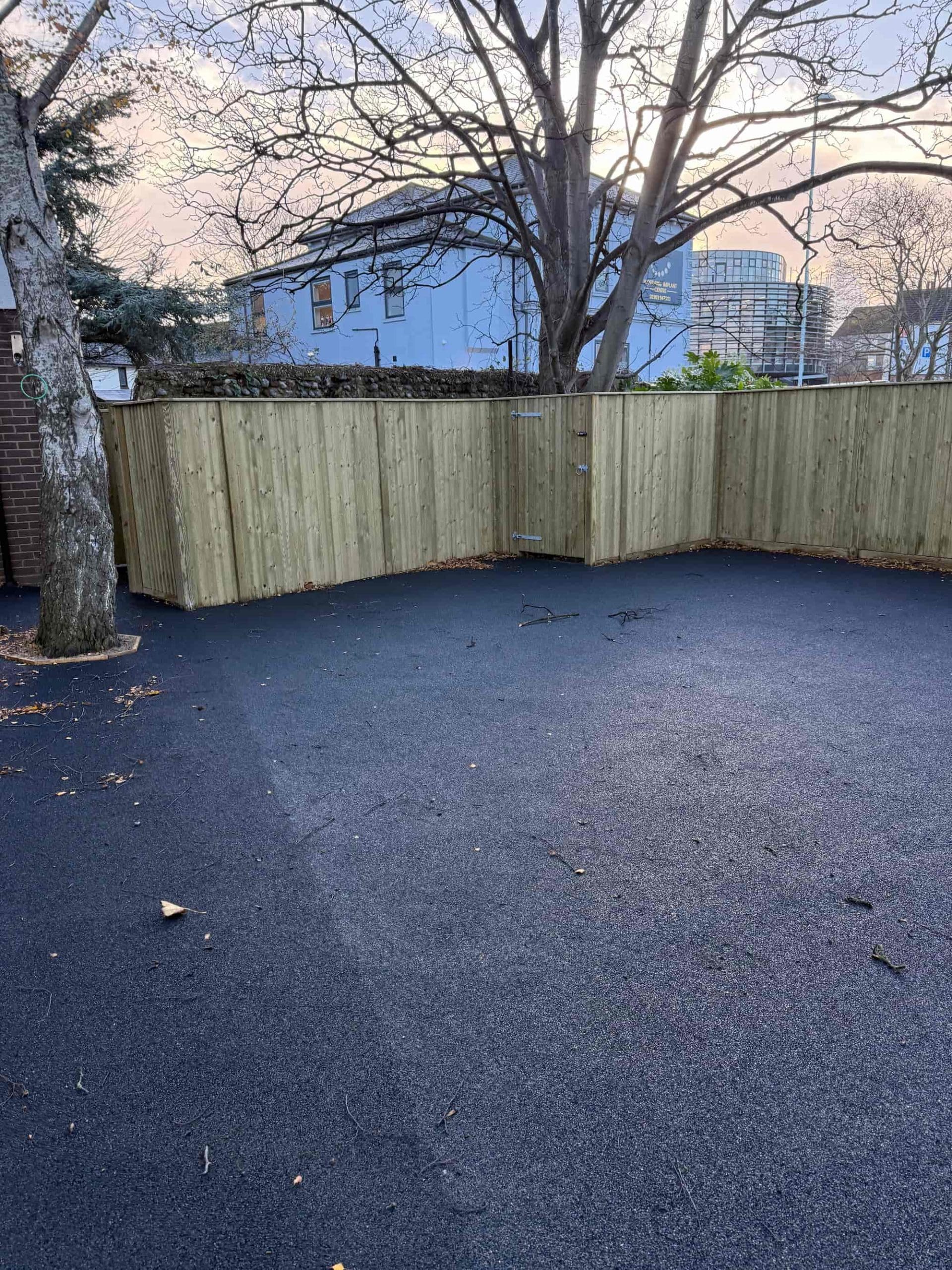 A fenced outdoor area with new wooden fencing and fresh black asphalt ground; a leafless tree stands on the left, and buildings are visible in the background under a cloudy sky.