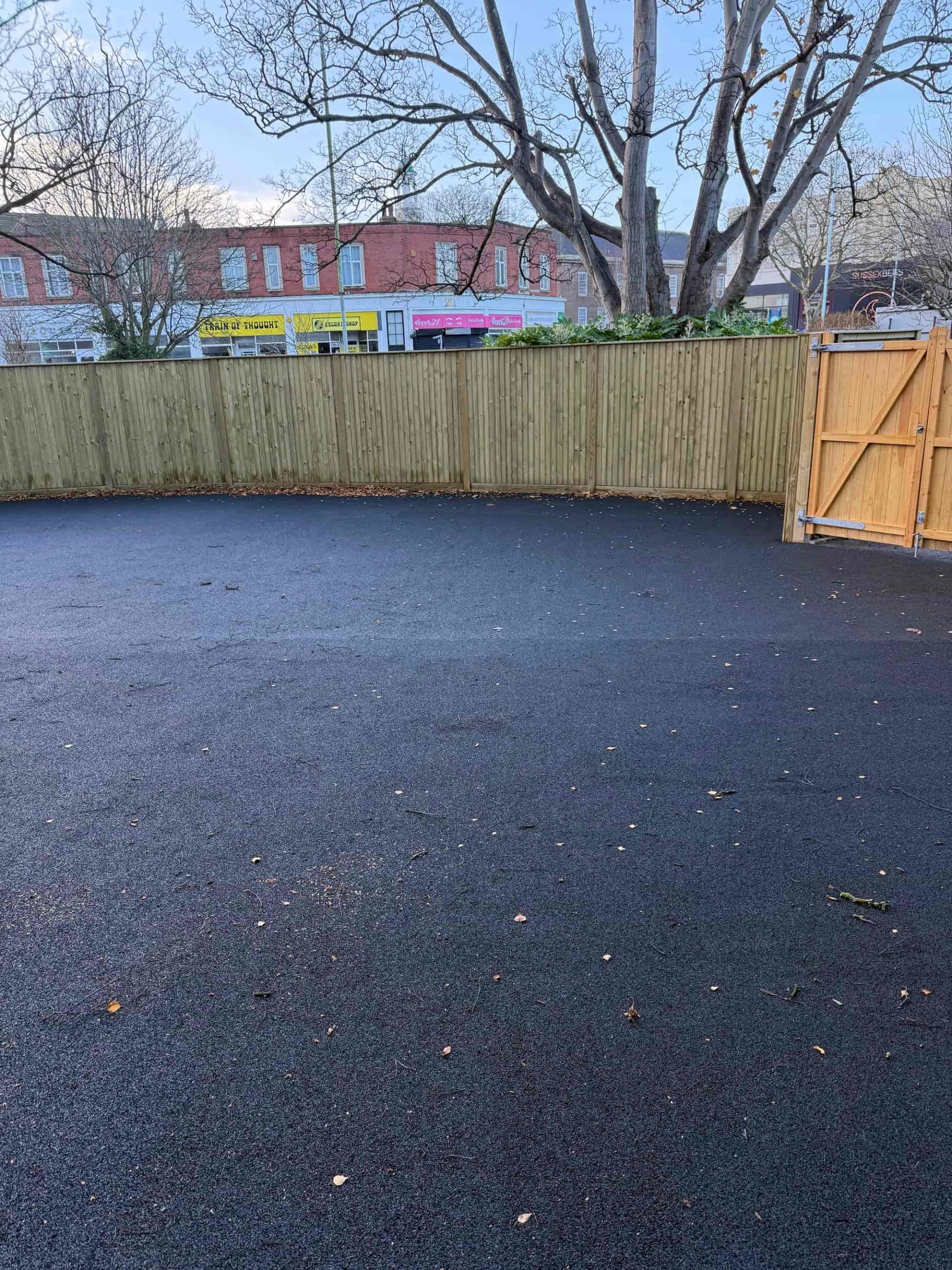 A freshly paved asphalt area bordered by a wooden fence with an open gate. Leafless trees and several shopfronts with colorful signs are visible in the background under a cloudy sky.