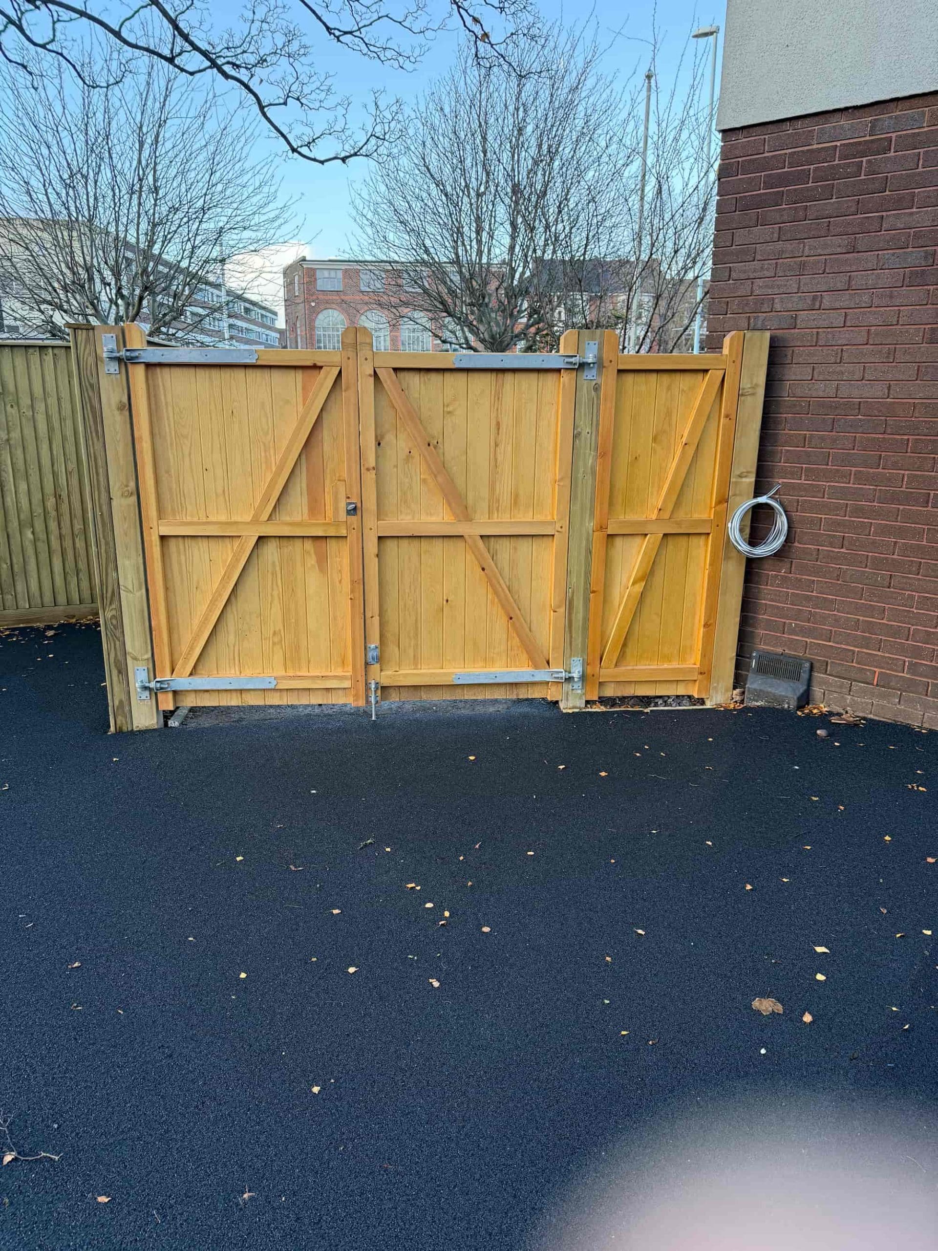 A wooden double gate with metal hinges stands closed next to a brick wall on black pavement. Leafless trees and a building are visible in the background, with a coiled pipe hanging on the wall.