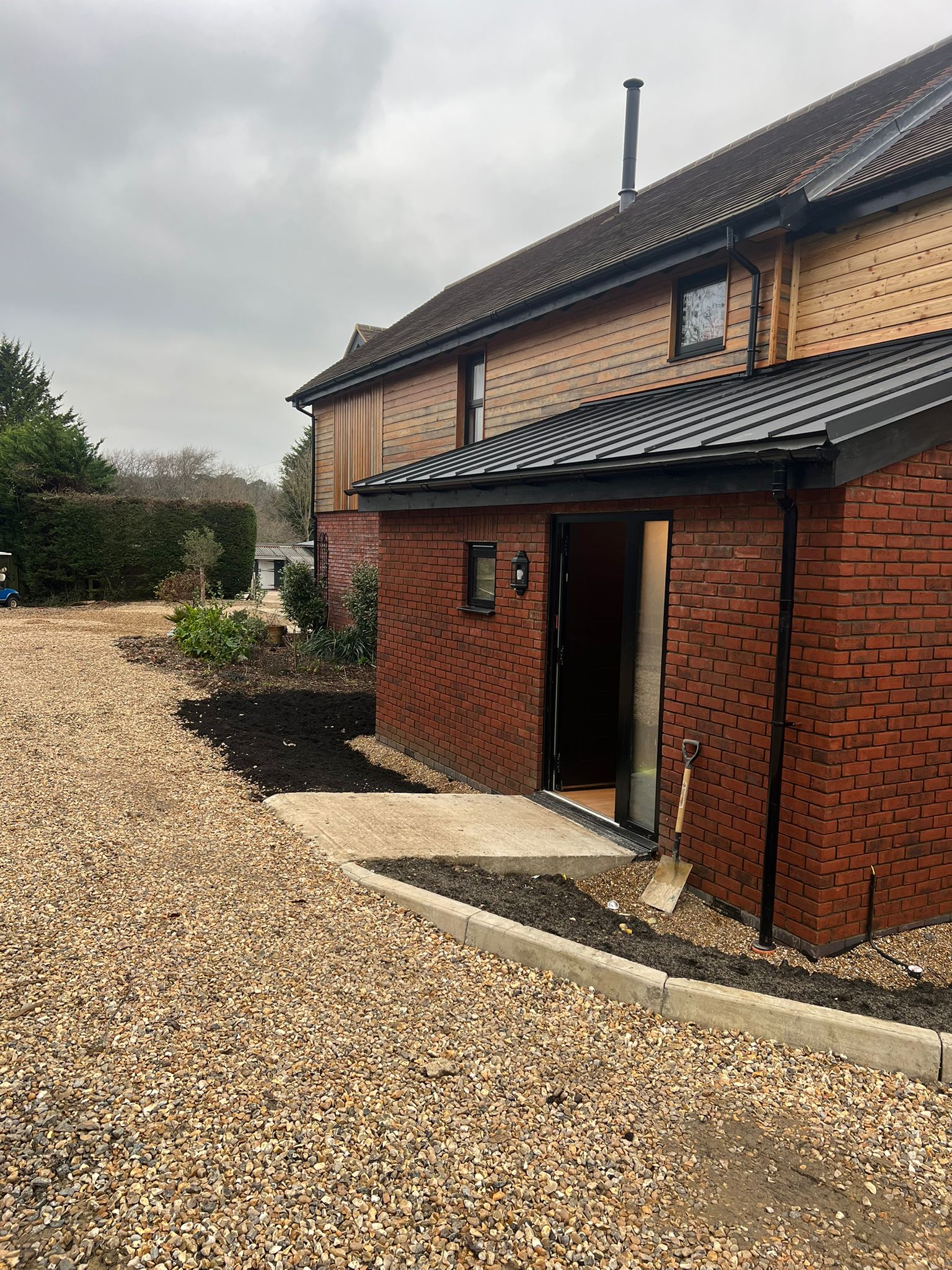 A modern brick and wood house with an open door, gravel driveway, and a small garden area. A shovel is leaning against the wall near a newly laid concrete path. The sky is cloudy.