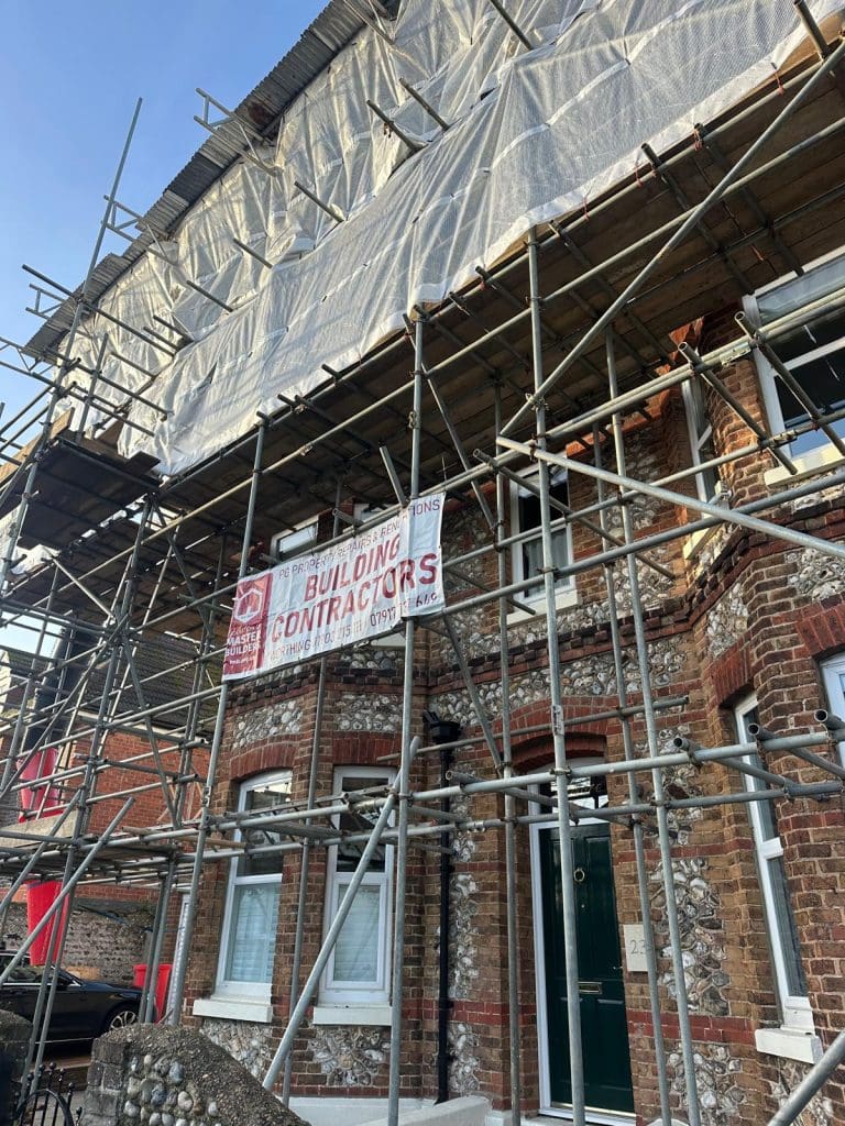 A brick house covered in scaffolding with a tarpaulin overhead. A sign for a building contractor is attached to the scaffolding above the door and windows.