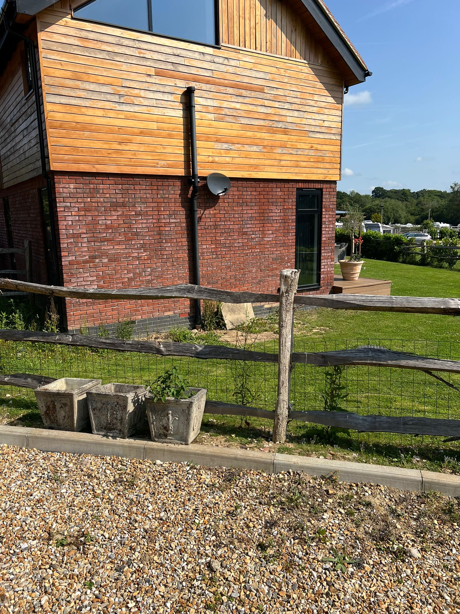 A modern house with wooden siding and red brick walls stands behind a wooden fence. There are three concrete planters in front, a green lawn, and a clear blue sky in the background.
