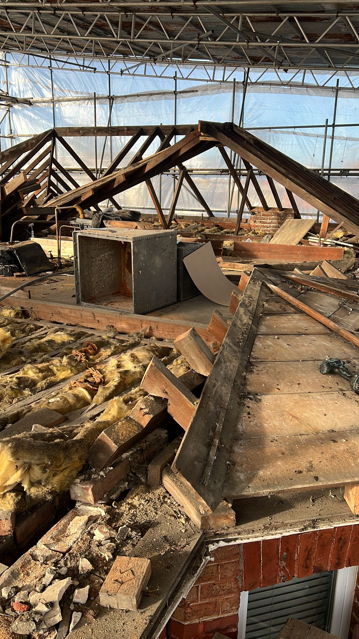 A partially demolished roof with exposed wooden beams, insulation, bricks, and debris scattered across the floor. The area is surrounded by scaffolding and translucent plastic sheeting, indicating ongoing construction work.