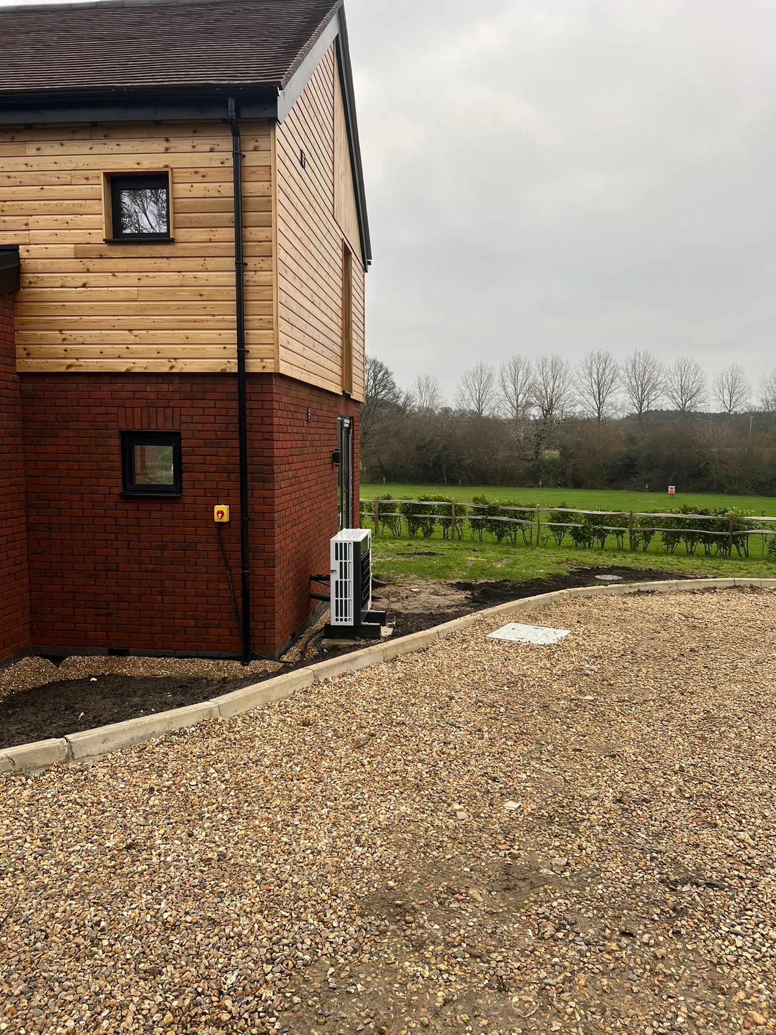 A corner of a modern brick and wood house with an air conditioning unit outside, gravel driveway, and a grassy field with picnic benches and trees in the background under a cloudy sky.