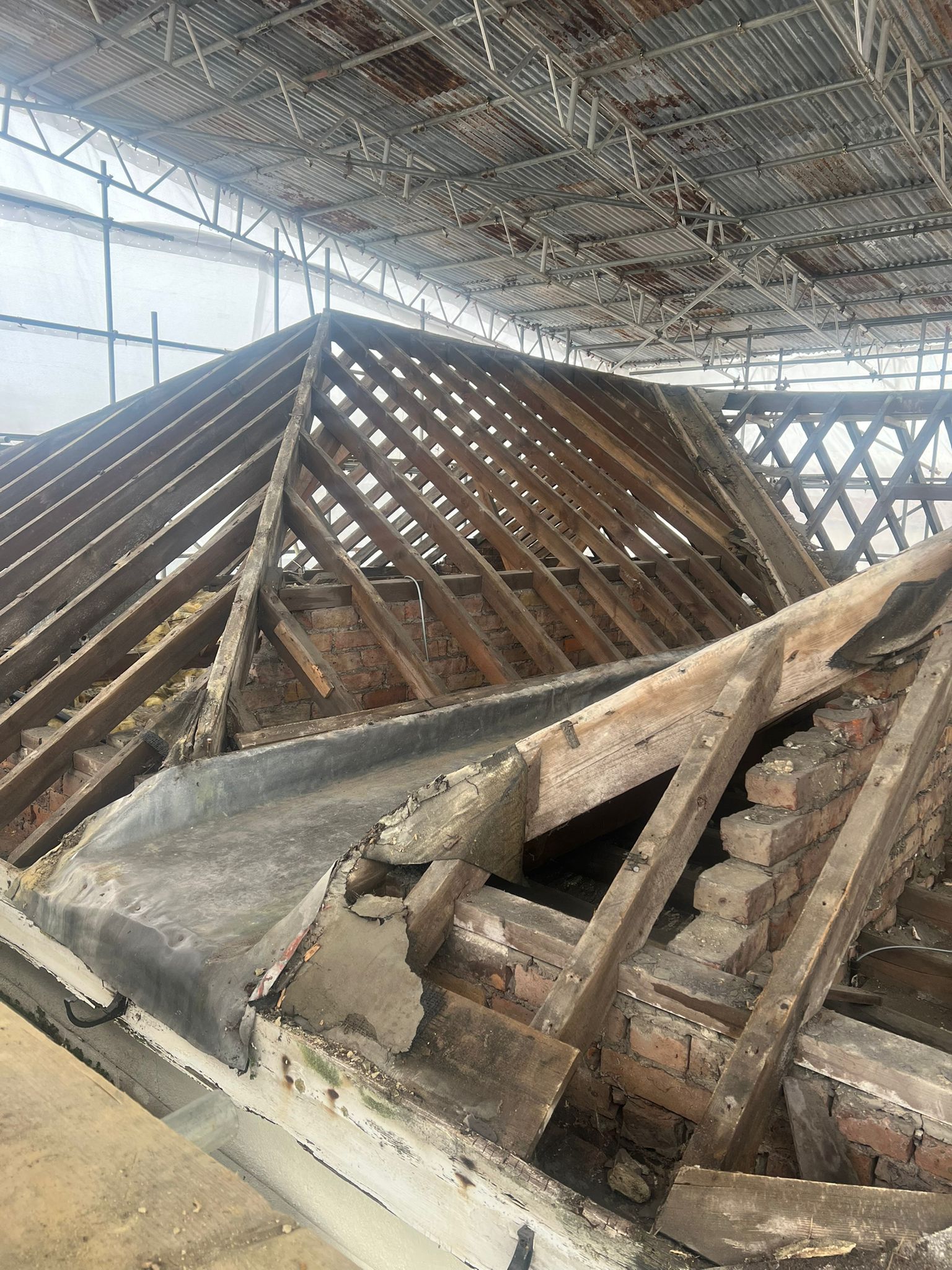 A damaged roof with exposed wooden beams and bricks, showing significant decay and missing tiles, under a metal scaffolding structure for repairs or restoration.