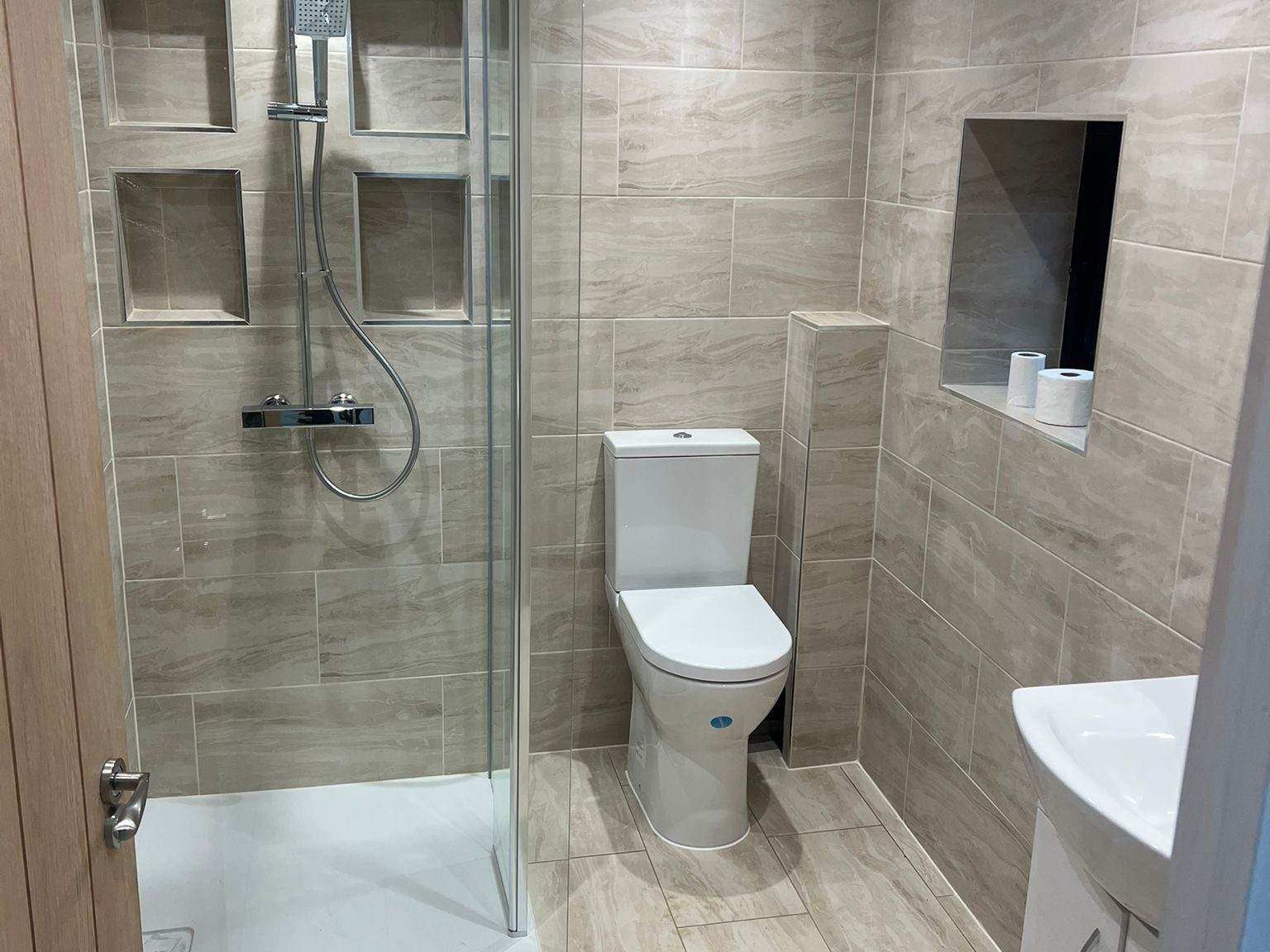 Modern bathroom with beige tiles, a glass-enclosed shower with built-in shelves, a toilet, a small sink with a cabinet, and a recessed mirror above the sink. The room is well-lit with ceiling lights.