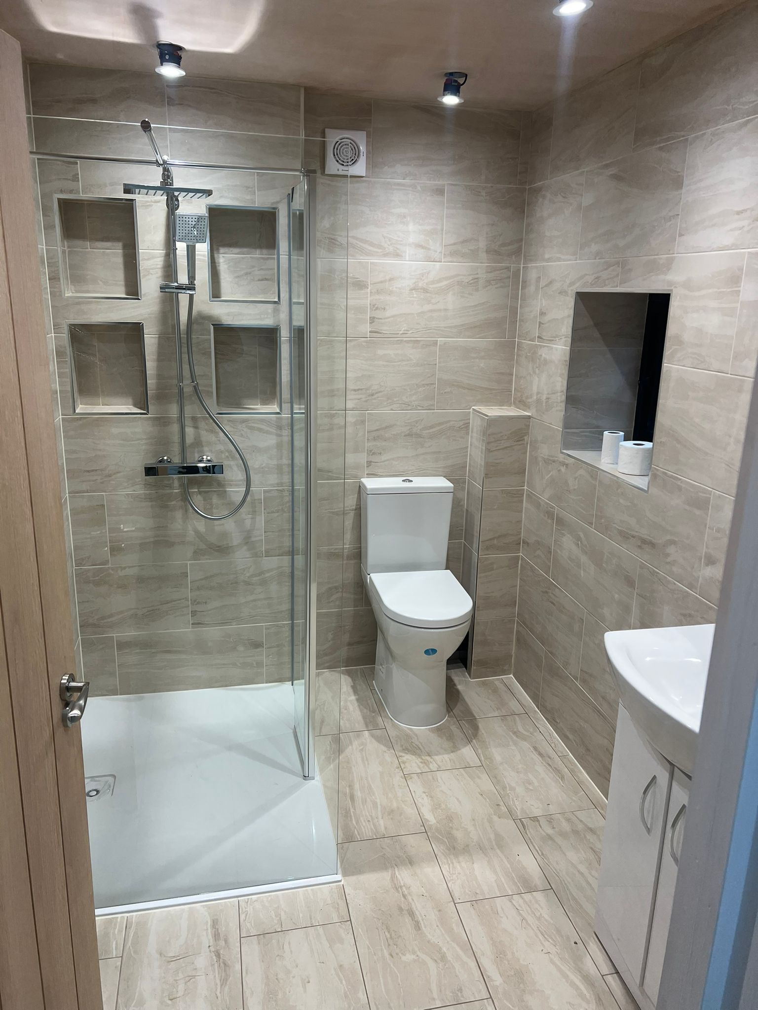 Modern bathroom with beige tiles, a glass-enclosed shower with built-in shelves, a toilet, a small sink with a cabinet, and a recessed mirror above the sink. The room is well-lit with ceiling lights.