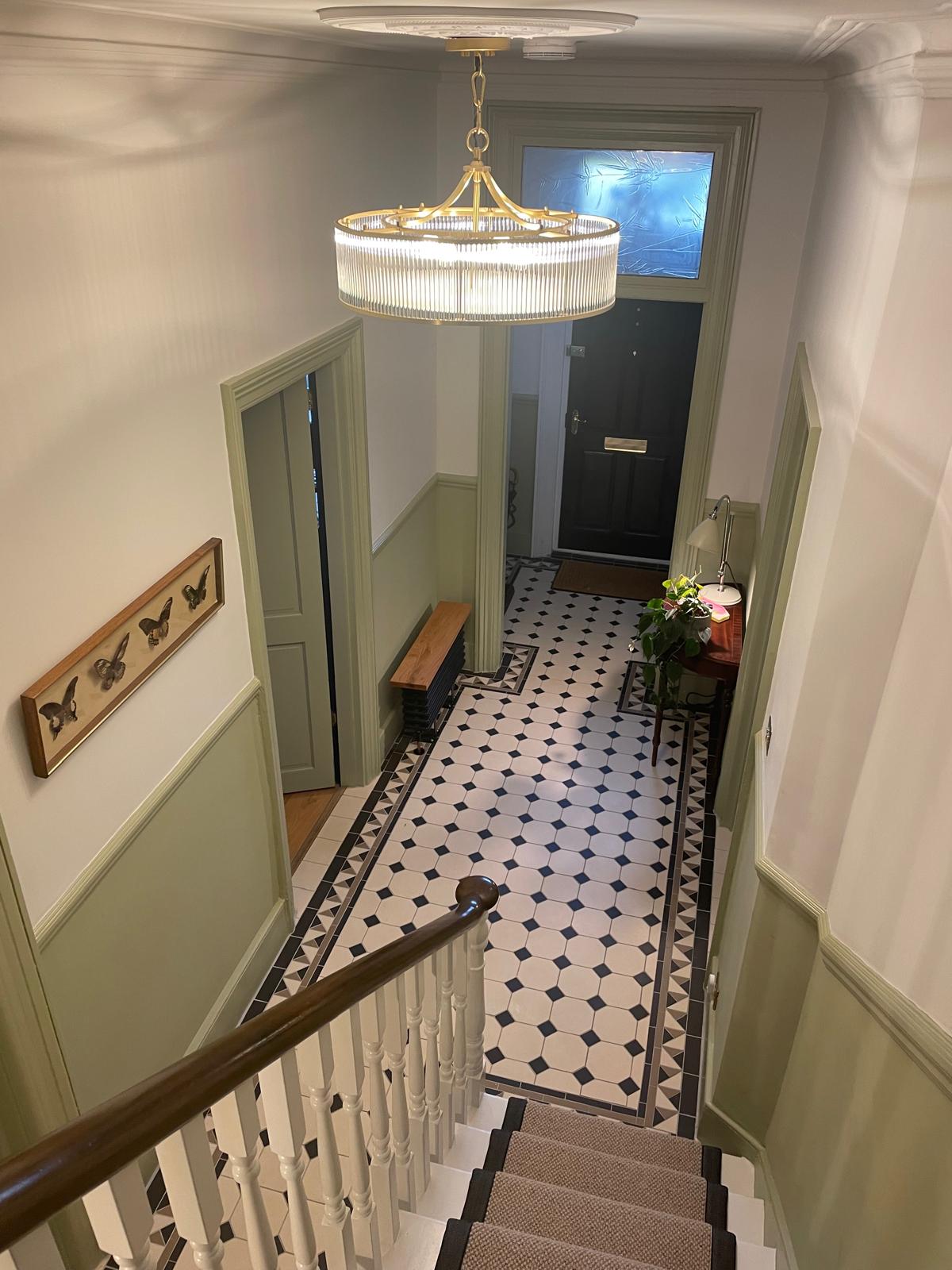 View from the top of a staircase looking down to a hallway with patterned tile floor, green trim, a black front door, a hanging light fixture, and a wooden bench with a plant and framed butterfly art on the wall.