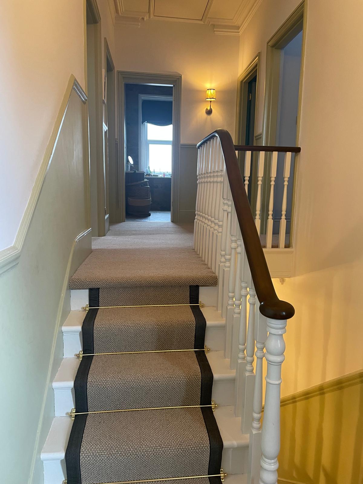 A staircase with a beige carpet runner and brass stair rods leads to a hallway with light walls, green door frames, and a wooden railing, illuminated by a wall sconce near the end.