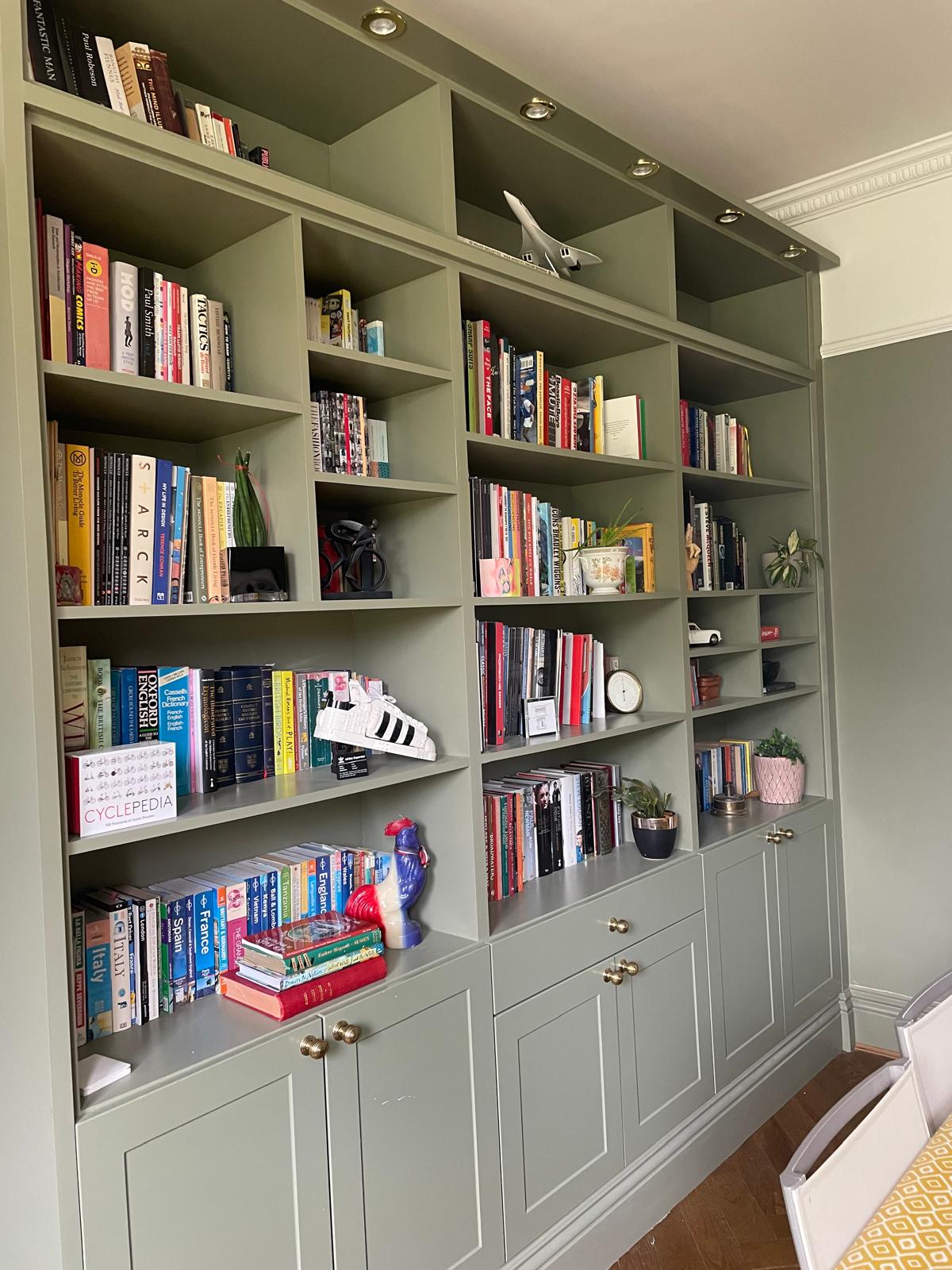 A green built-in bookshelf filled with colorful books, potted plants, ornaments, a clock, a toy rocket, a model airplane, and a pair of white sneakers on the middle shelf. The lower part has closed cabinets.
