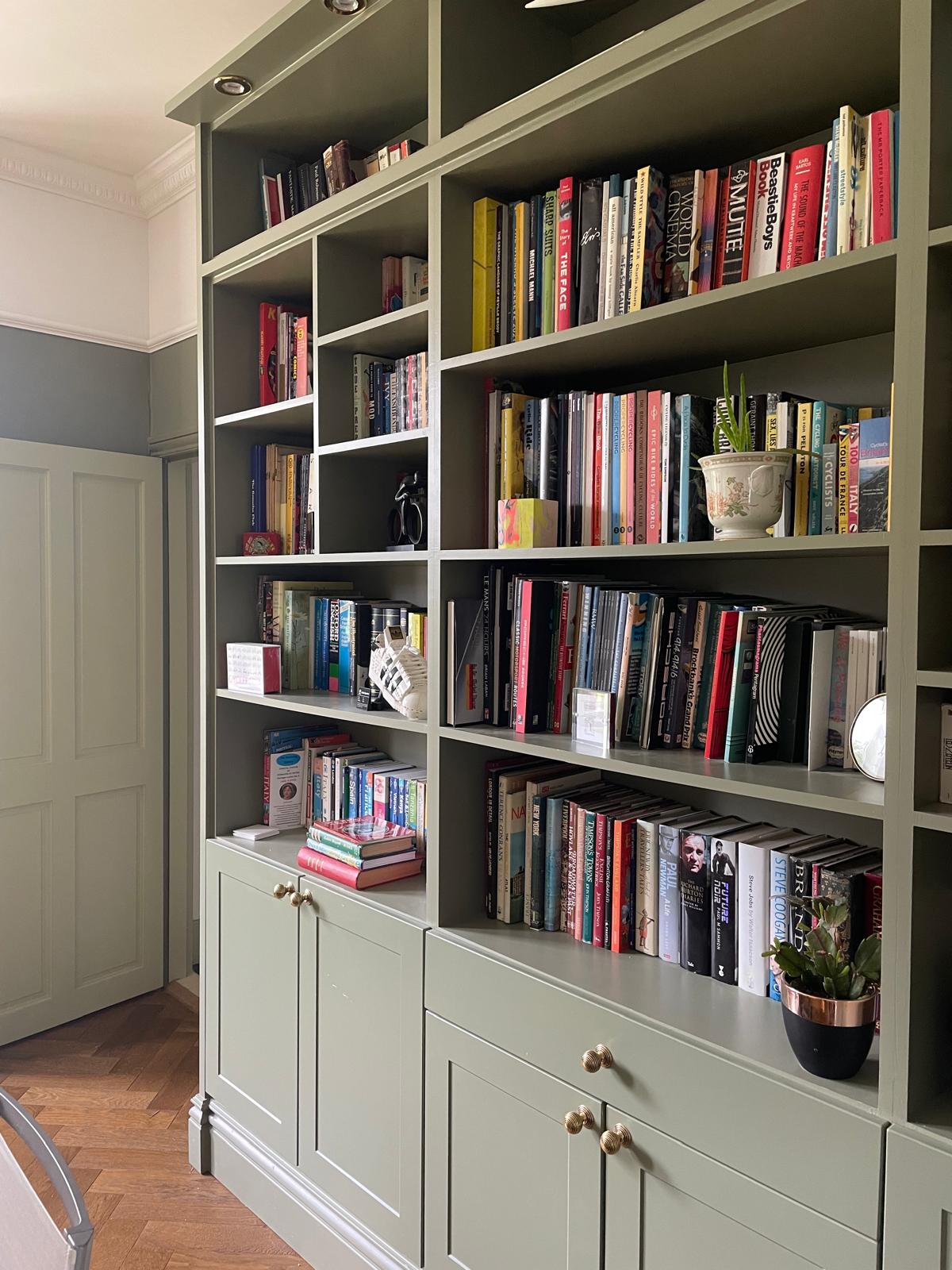 Built-in green shelves and cabinets filled with neatly arranged books, a potted plant, framed photo, and decorative items. Wooden herringbone floor and soft, natural light brighten the room.