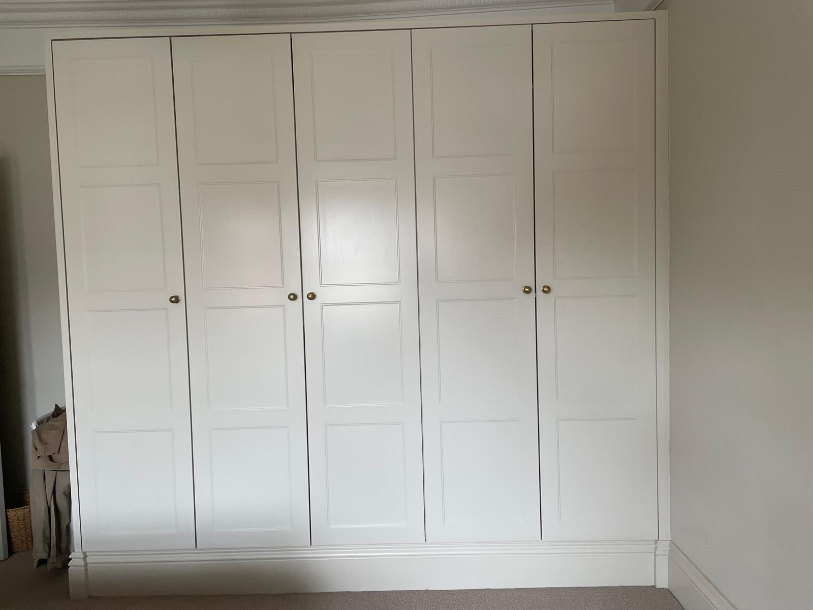 A built-in wardrobe with six white paneled doors, each featuring a small round knob, set against a light-colored wall in a carpeted room. A basket and some items are visible in the left corner.