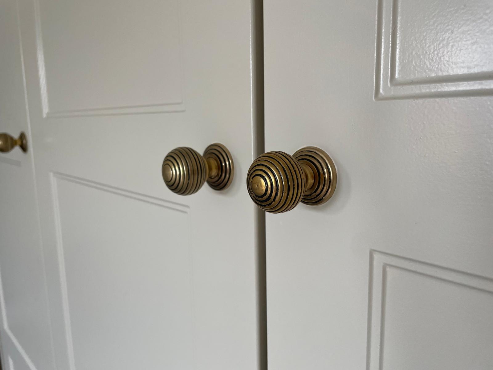 Close-up of white cabinet doors with gold, round, ridged handles. The doors have simple rectangular panel detailing and the handles are centered on the frame, casting soft shadows.